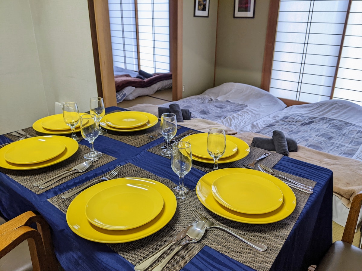 A dining table is set with yellow plates and glassware, accompanied by polished cutlery. In the background, traditional tatami mats are visible, featuring spacious beds arranged with soft bedding and decorative pillows. Light filters through paper sliding doors, creating a serene dining atmosphere.