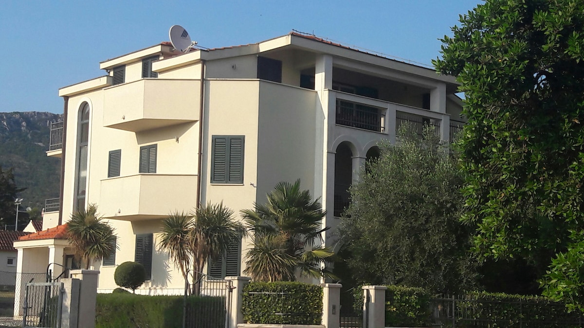 The exterior of a multi-story building is shown, featuring a combination of modern architectural elements and lush greenery. Balconies can be seen, along with palm trees and neatly trimmed shrubs surrounding the property. A satellite dish is visible on the roof.