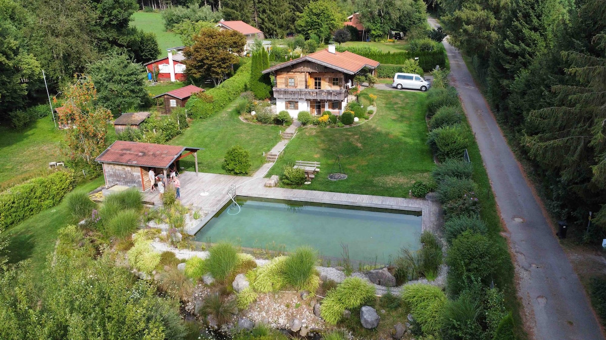 An aerial view captures a spacious wooden house surrounded by lush greenery. A natural pond is positioned nearby, reflecting the surrounding flora. Several outdoor seating areas are visible, along with a pathway leading to the nearby road, creating a serene setting in a quiet environment.