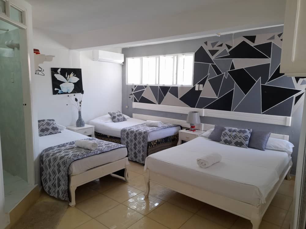 A bright room features three beds, two with white frames and one with a sleek design. A geometric wall mural adds visual interest, while light streams through a row of windows. Nightstands flank each bed, providing bedside greenery and soft linens.