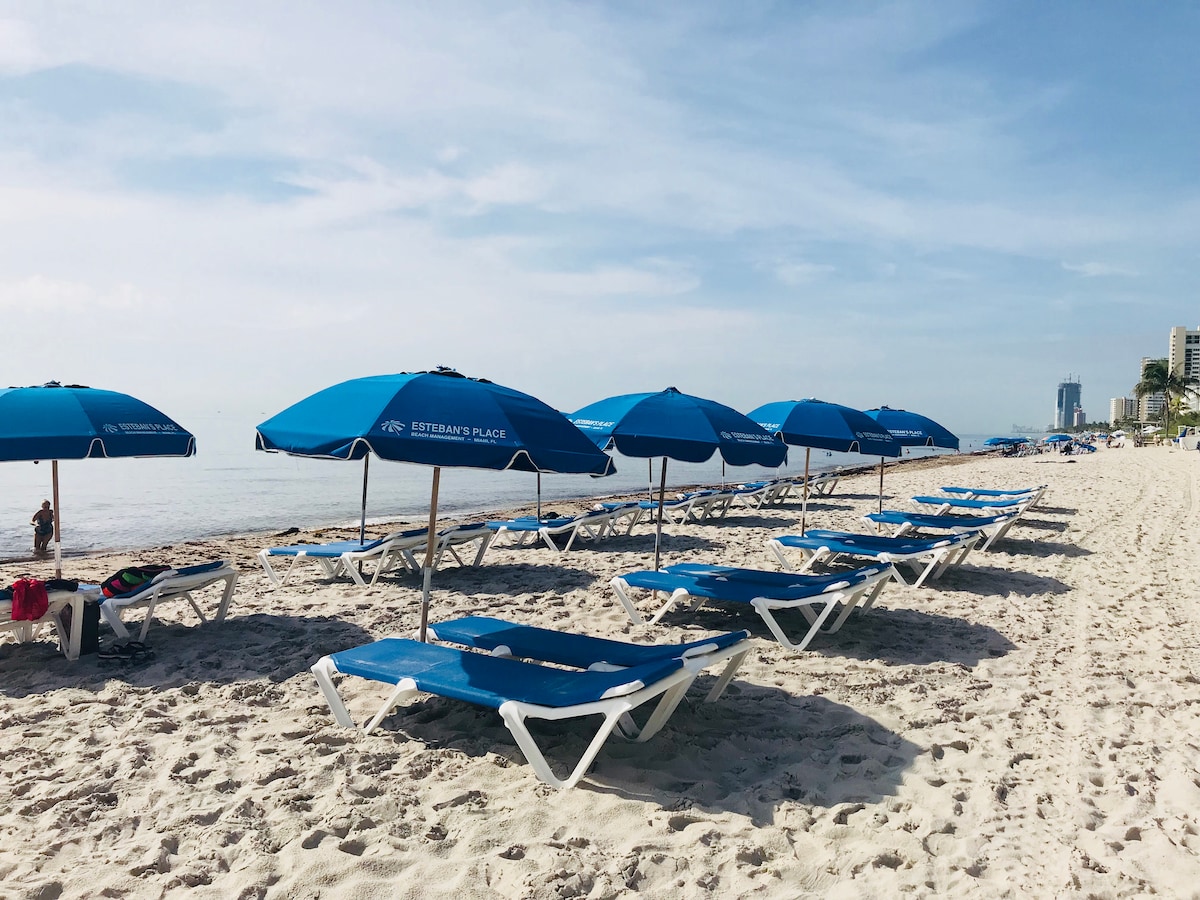A serene beach scene features numerous blue lounge chairs paired with matching umbrellas, arranged neatly along the soft, white sand. The calm ocean can be seen in the distance, with a clear sky providing a peaceful backdrop.