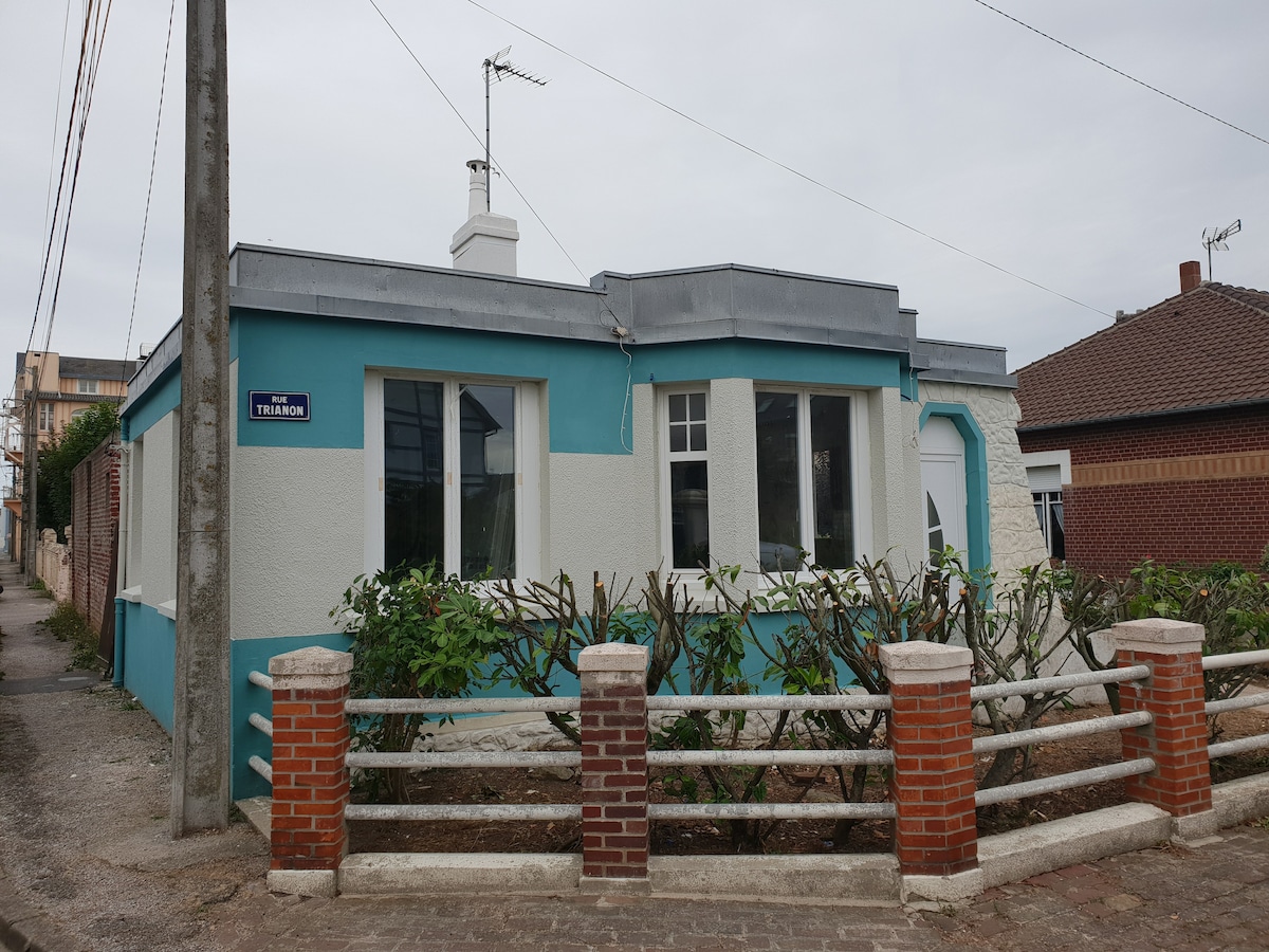 A charming single-story house is presented with a light blue trim and bright white exterior. Large windows allow natural light to fill the interior, while a low fence and greenery frame the property, contributing to a welcoming entrance in a residential neighborhood.
