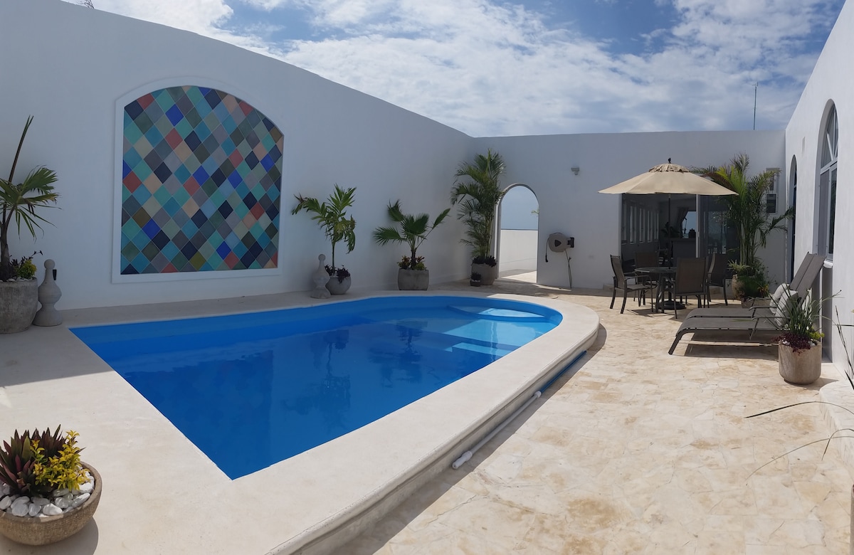 A private pool area features a bright blue swimming pool surrounded by a tiled deck. Lush potted plants are placed strategically, and comfortable lounge chairs are arranged nearby. A shaded dining area with a light-colored canopy is visible, offering an inviting space for outdoor meals.