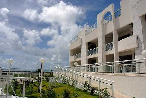 A modern building with a light-colored facade is shown, featuring balconies and a manicured lawn with greenery. Pathways lined with lamp posts lead to the entrance, under a partly cloudy sky, suggesting a bright and airy environment.