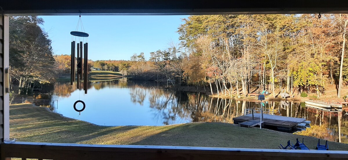 A serene view of Indian Rock Lake is framed by large windows. The calm water reflects the bright blue sky and surrounding trees, showcasing vibrant autumn colors. A fishing dock is visible along the shore, enhancing the peaceful lakeside setting.