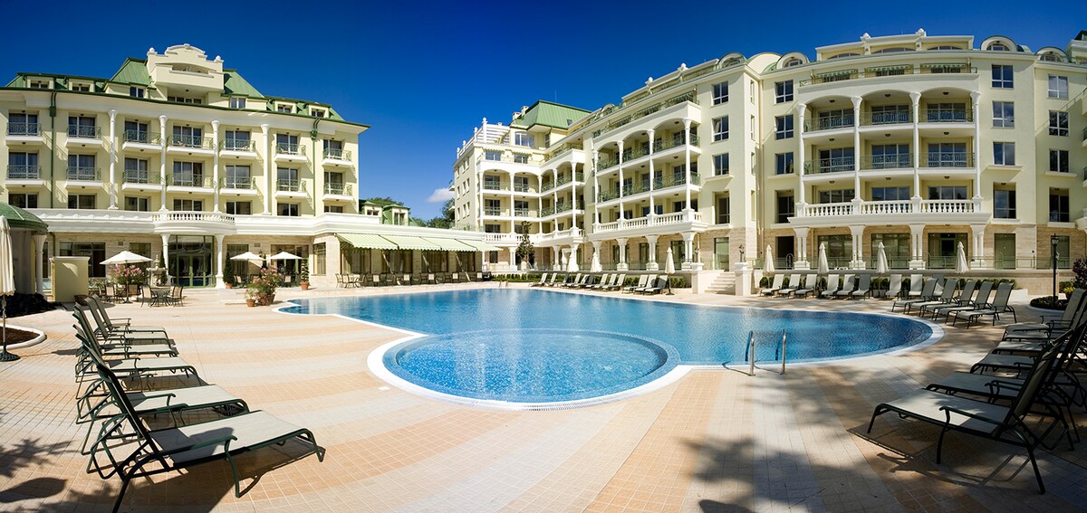 The spacious outdoor pool is surrounded by loungers, offering ample relaxation space. The three-story building features large balconies and green roofs, set against a clear blue sky. Sun umbrellas are placed at intervals, enhancing the inviting atmosphere of the pool area.