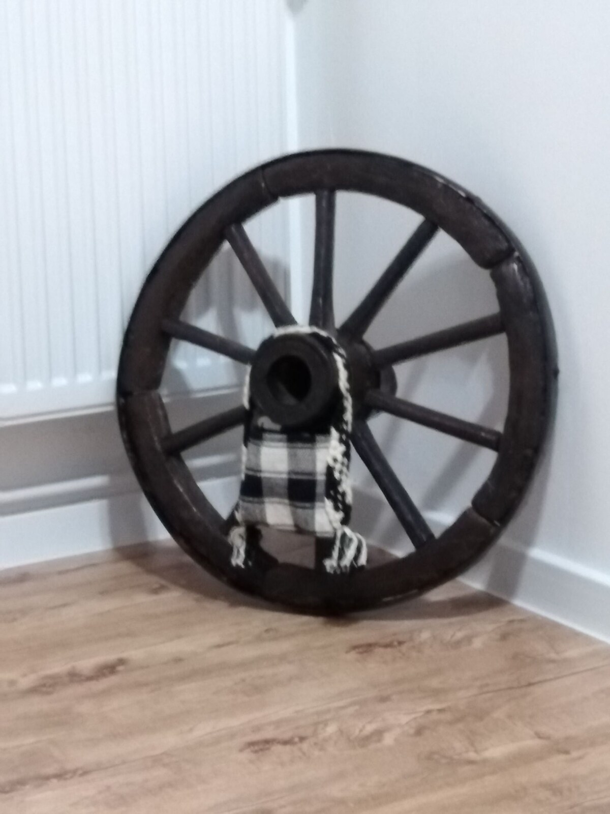 An antique wooden wheel is positioned in the corner of the room, showcasing eight spokes and a central hub. A black and white woven textile is draped through the wheel's center, adding a rustic touch to the modern decor. Light wood flooring complements the overall design.