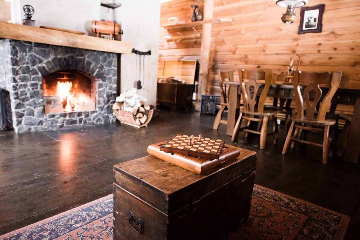 A warm living space features a stone fireplace with a crackling fire. A wooden table and chairs are arranged nearby, while a chess set is displayed on a vintage trunk in the foreground. Natural wood paneling complements the inviting atmosphere.