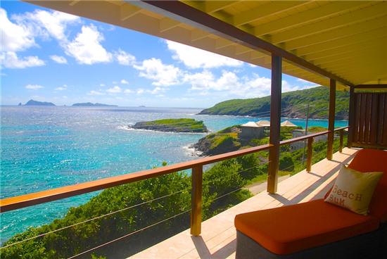 A wraparound balcony offers stunning ocean and island views, with turquoise waters and lush green hills visible in the distance. Outdoor seating is provided, allowing for a relaxing perspective of the coastal landscape under a bright blue sky.