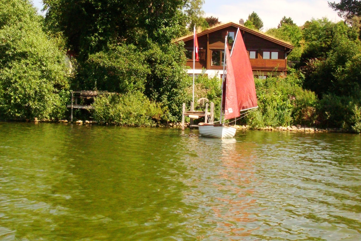A small sailing boat with a red sail is anchored near lush greenery, facing a brown wooden house framed by trees. The calm water reflects the surrounding foliage, creating a serene setting suitable for relaxation.