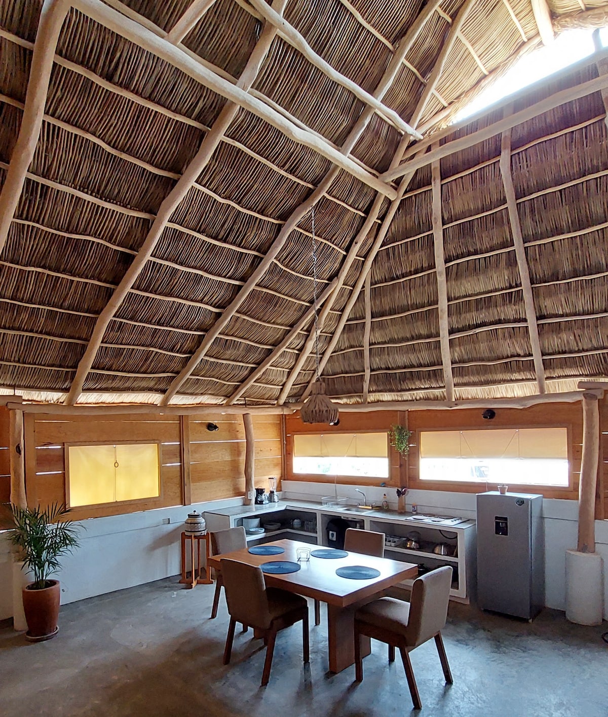 A spacious kitchen area features a high thatched ceiling and wood beams. A dining table is set for four, accompanied by comfortable chairs. A modern kitchen counter offers appliances and a large refrigerator, while sunlight filters through wide windows dressed in simple shades.