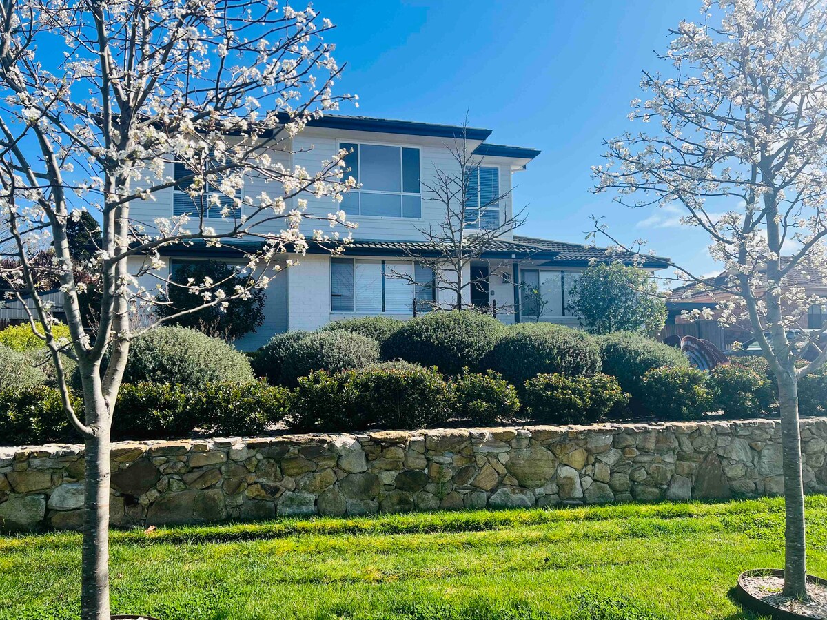 A two-story home is set against a clear blue sky, surrounded by blooming trees and manicured shrubs. A stone wall enhances the garden area, with a lush green lawn providing a vibrant foreground.