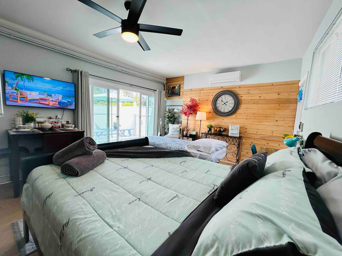 The sleeping area features a spacious bed with light green bedding and various cushions. A television is mounted on the wall, while a large window with sliding doors provides natural light, leading to the outdoor patio. A small table with decorative items complements the room's decor.