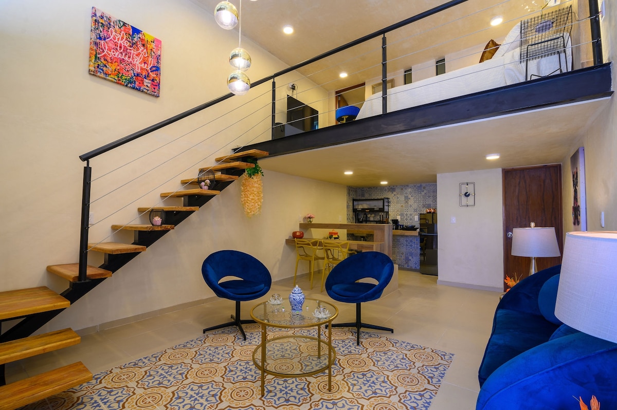 The spacious loft features a modern open layout with a staircase leading to the upper sleeping area. Two blue chairs are positioned around a round coffee table on a patterned rug, while pendant lights hang from the ceiling, illuminating the inviting space.