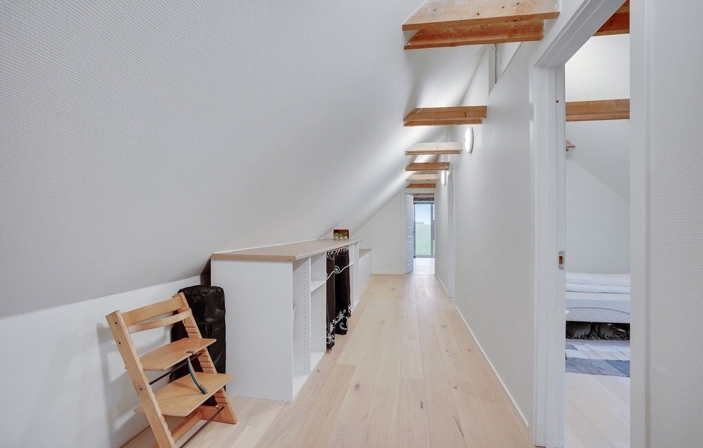 A well-lit hallway features a light-colored wooden floor and white walls. The ceiling is adorned with wooden beams, enhancing the spacious feel. A small, simple chair is positioned against one wall, with built-in shelving visible nearby. Natural light enters through a distant window.