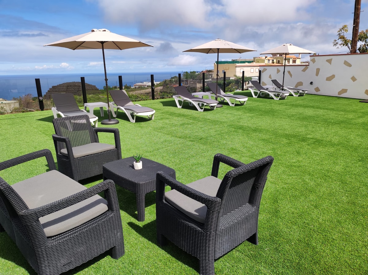An outdoor lounge area is seen featuring gray wicker seating with cushions and a low table. Four sun loungers with adjustable backs are arranged under two umbrellas. A lush green lawn stretches across the area, with views of the sea visible in the background.
