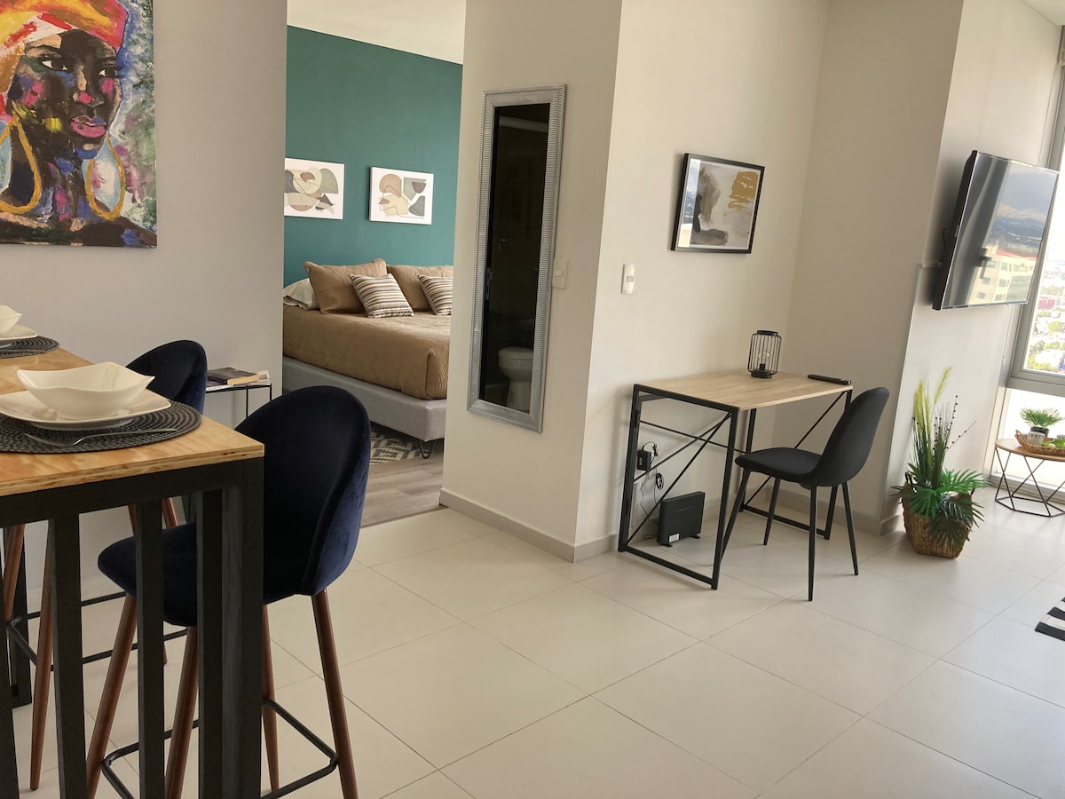 A modern dining area features a high-top table with four bar stools, set for a meal. In the background, a cozy sleeping space with neutral bedding and artwork is visible. A desk is positioned beside a large window, providing a workspace with natural light.