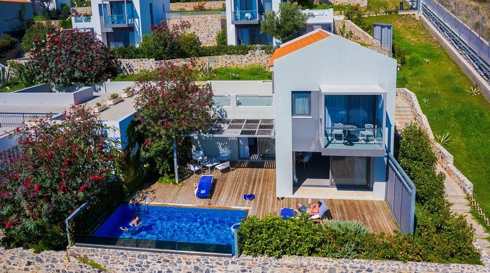 The villa is set against a scenic backdrop, featuring a private pool surrounded by a wooden deck. Lounge chairs are arranged near the pool, and a shaded balcony overlooks the serene landscape. Lush greenery and flowering plants enhance the outdoor space.