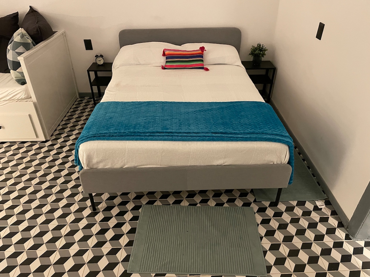 The sleeping area features a spacious bed with a light-colored mattress and decorative pillow. A vibrant blue blanket adds a pop of color. Flanking the bed are two nightstands. The floor showcases a geometric tile pattern, complemented by a soft area rug.