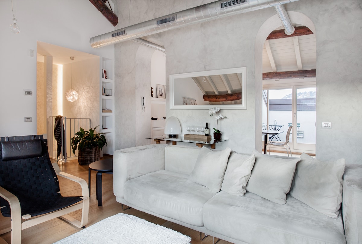 A spacious living area features a light-colored sectional sofa and a sleek black lounge chair. Exposed log beams add character to the ceiling, while a modern mirror reflects natural light. An entrance to a balcony is visible in the background, enhancing the open feel of the space.