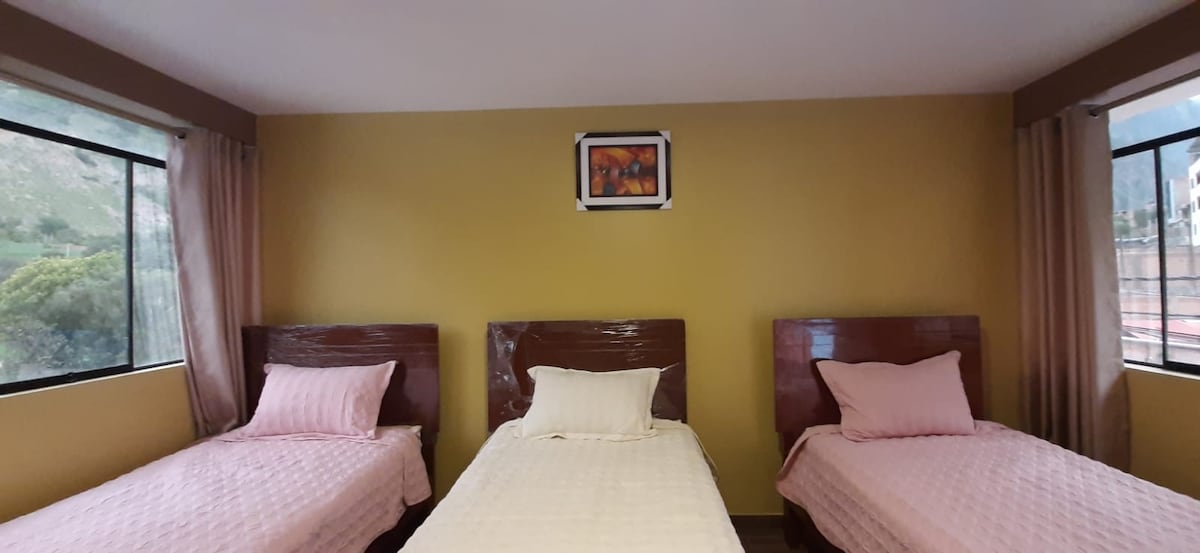 A room featuring three triple beds, each dressed in soft pink linens. Light streams in through large windows adorned with sheer curtains, while a colorful wall art piece adds a touch of character to the yellow walls.