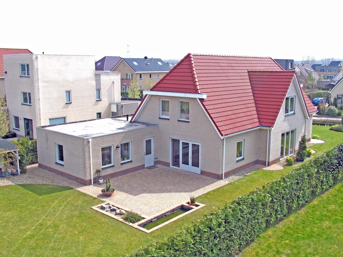 An aerial view showcases a spacious villa with a distinctive red roof, surrounded by well-maintained gardens and pathways. The exterior features a mix of stone and light-colored walls, with a patio area visible in the foreground, providing a glimpse of outdoor living space.