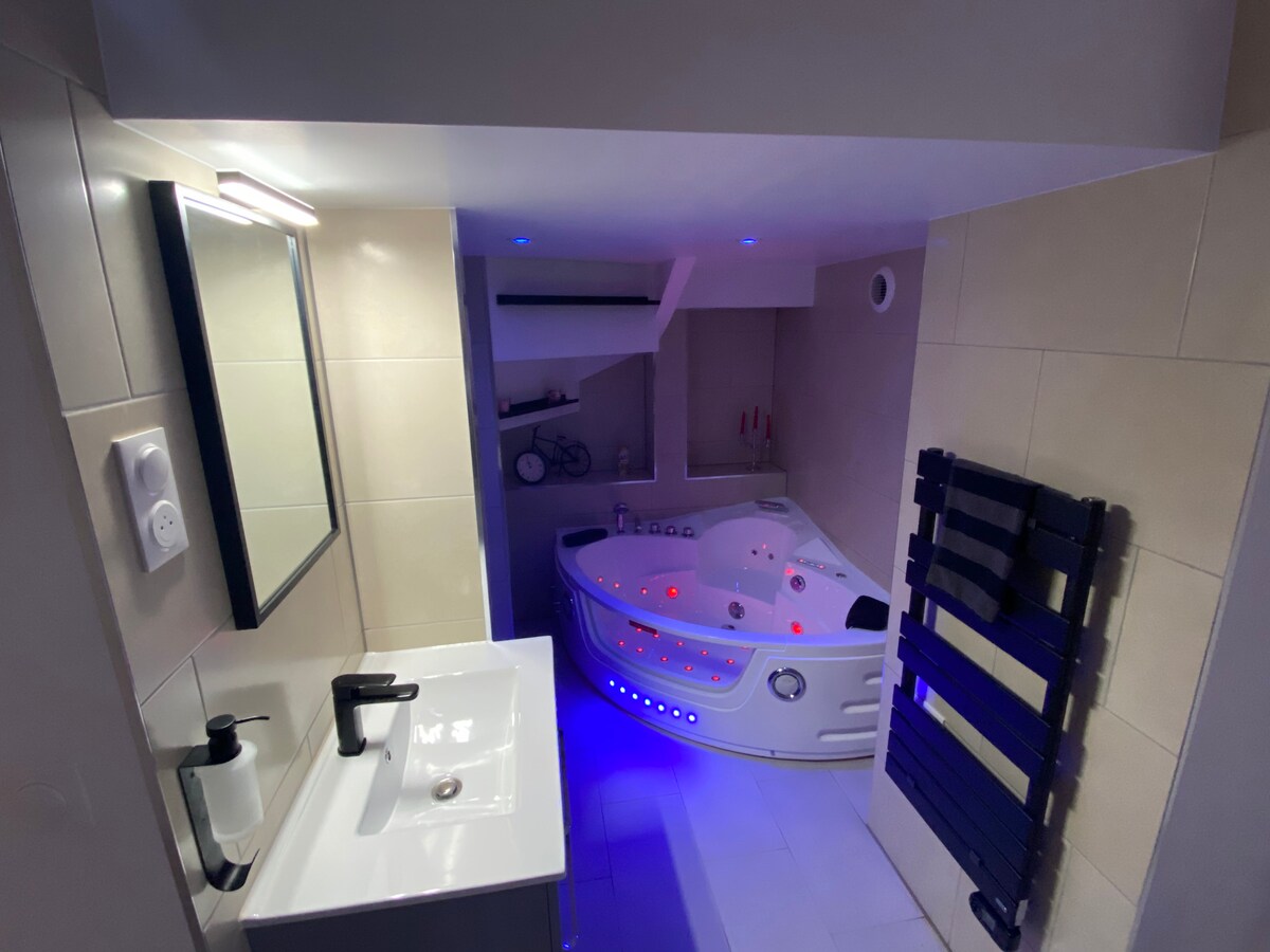 The bathroom features sleek, modern fixtures with a spacious washbasin and a large mirror. A luxurious whirlpool bathtub is illuminated with ambient lighting, creating a relaxing atmosphere, while a heated towel rail is positioned nearby.
