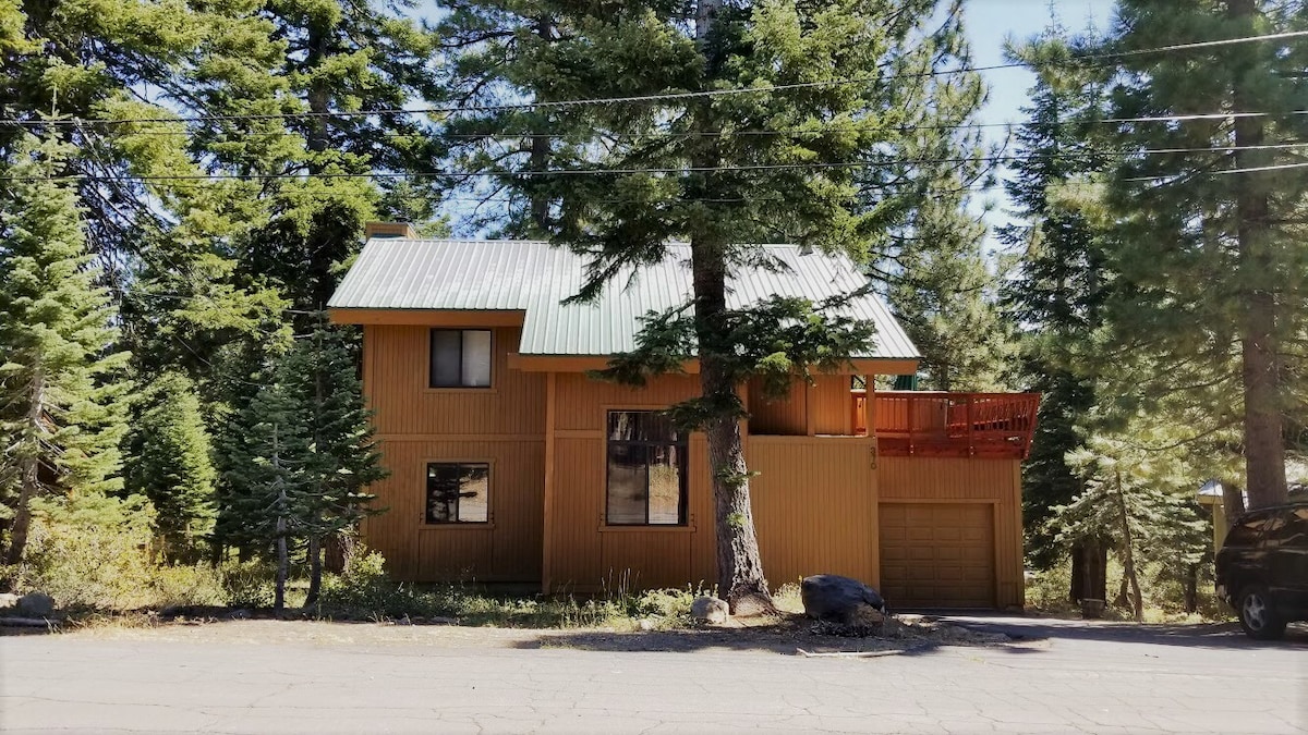 A multi-level cabin exterior is surrounded by tall trees, showcasing a wood-paneled façade and a green metal roof. The property features an upper deck accessed by stairs, with large windows allowing natural light to enter. A driveway leads to the garage, partially visible at ground level.
