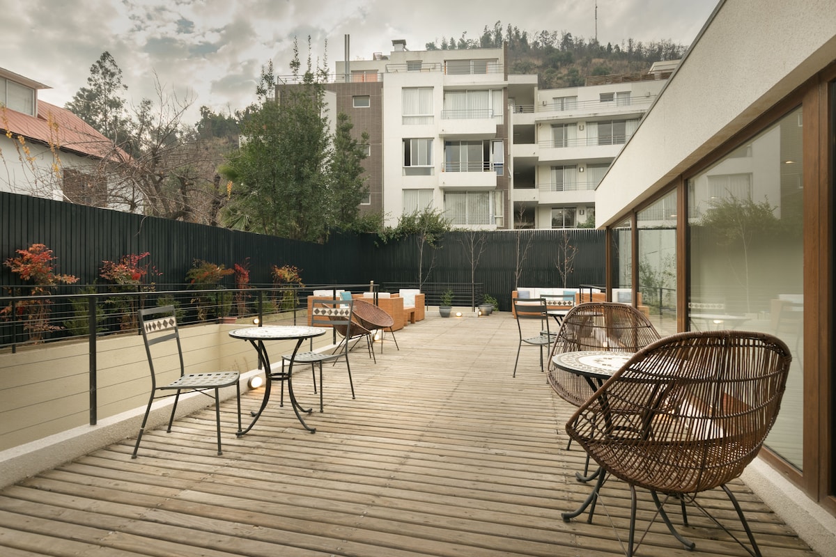 A spacious outdoor terrace is presented with wooden flooring and metal railings. Several seating arrangements, including round tables and comfortable wicker chairs, are visible. Lush greenery surrounds the area, providing a serene ambiance in a city environment.