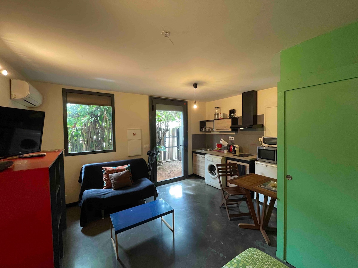 An open living space showcases a cozy sofa and a dining table. A kitchenette is equipped with modern appliances, including a washing machine. Natural light filters through the windows, illuminating the room which opens to a private garden area.