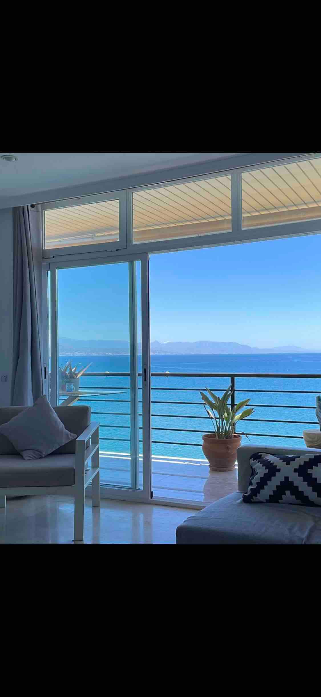 A bright living area features large glass doors that open to a balcony, showcasing an unobstructed view of the Mediterranean Sea and distant mountains. Potted plants add a touch of greenery, complementing the light-colored furnishings.