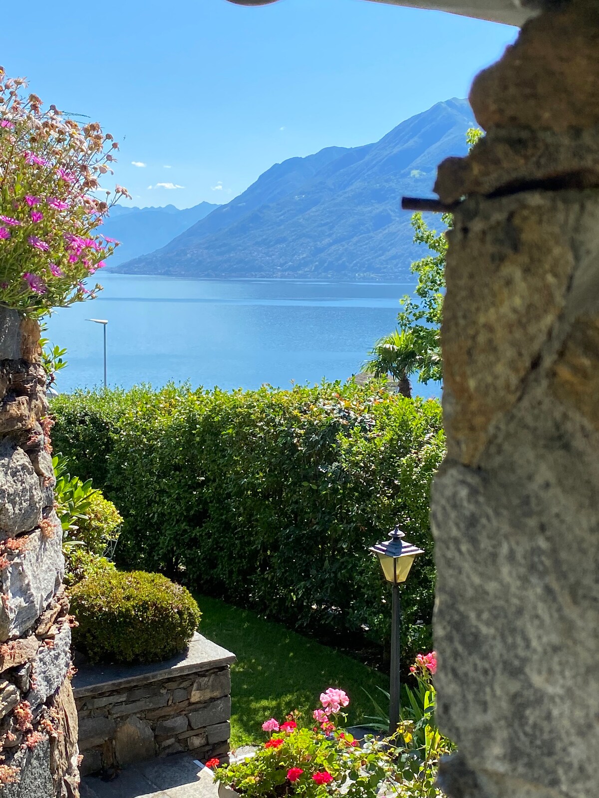 A serene view of the Lago Maggiore is framed by a stone wall and lush greenery. Colorful flowers bloom above and a lamp post is visible in the garden, adding charm. The distant mountains and clear blue sky reflect upon the water, creating a peaceful natural setting.