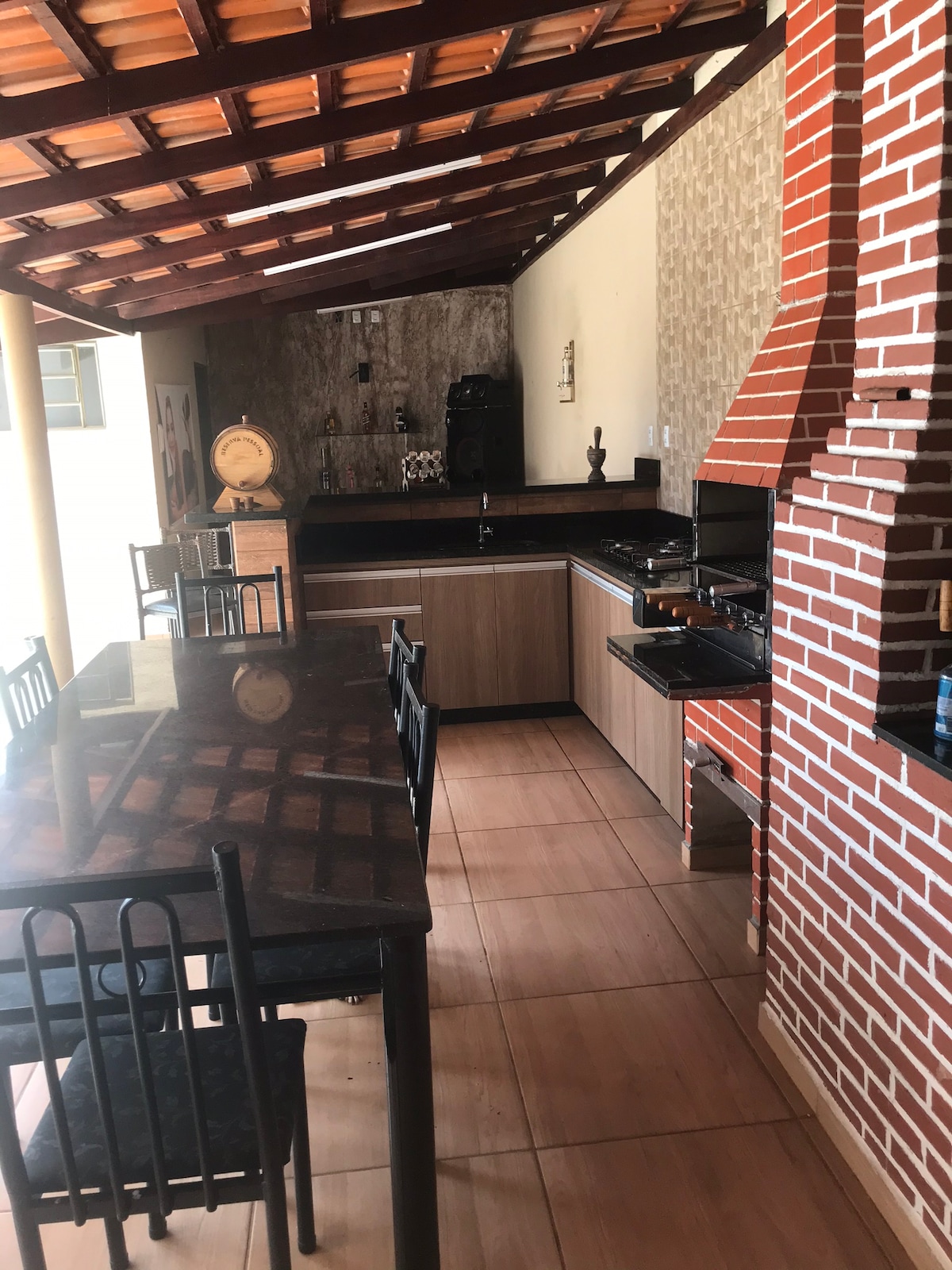 A spacious kitchen area features a long, dark countertop with several black chairs lined up. A brick grill setup is visible in the corner, complemented by modern appliances. Light filters in from the ceiling, highlighting the patterned wall and sleek cabinetry.