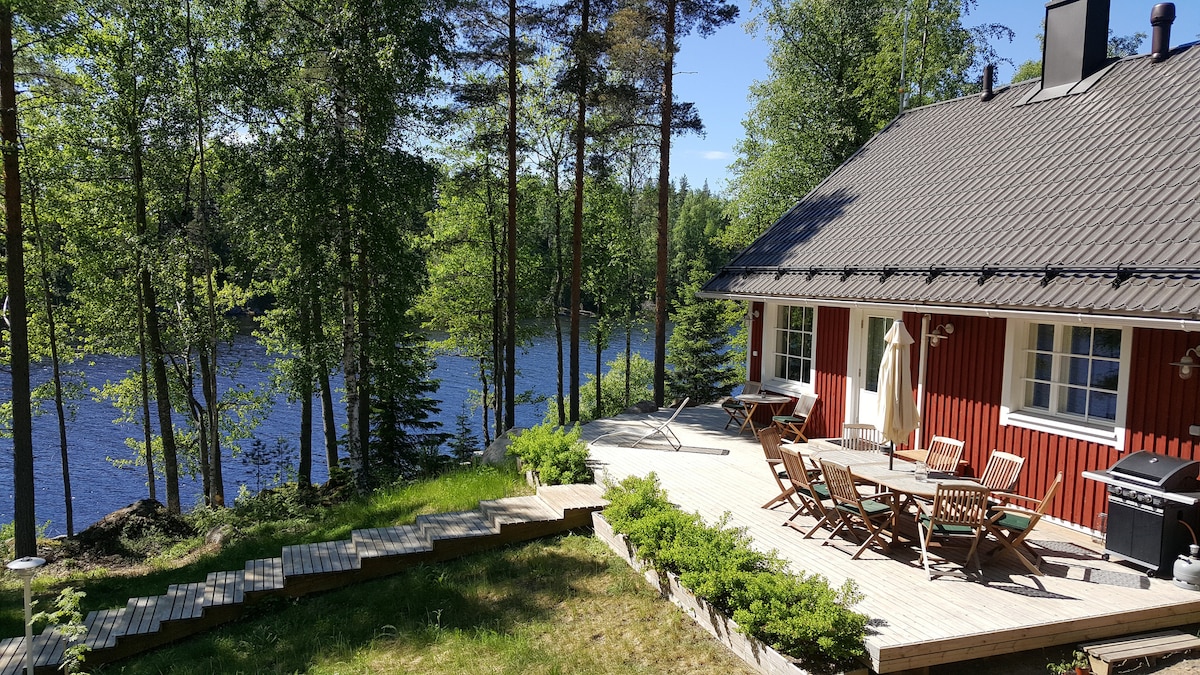 A red cottage is nestled among trees beside a lake. A wooden deck features a dining table surrounded by chairs, and a barbecue grill is visible. A grassy area leads to stairs descending towards the water, providing a serene view of the lake and surrounding nature.