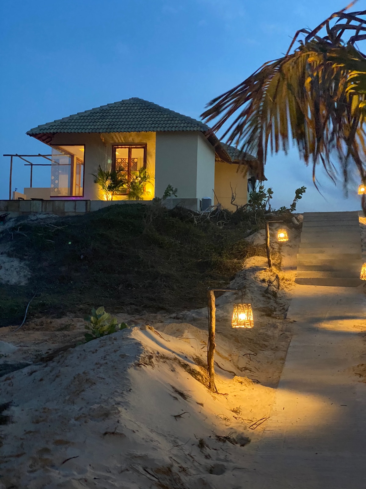 A pathway illuminated by lanterns leads to a charming bungalow, surrounded by sand and vegetation. The bungalow features a tile roof and large windows that reflect warm light, set against a dusky sky to create an inviting atmosphere.