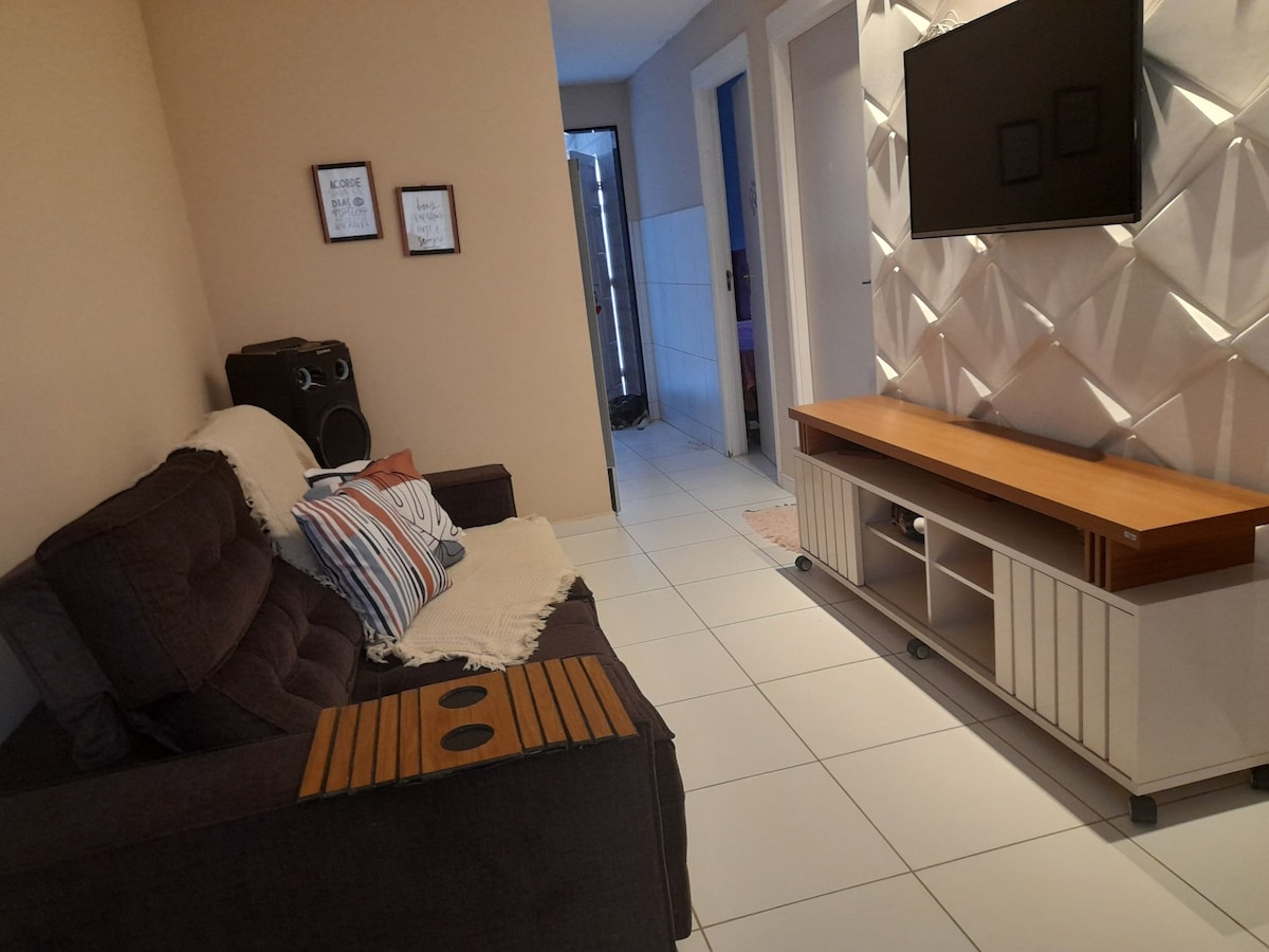 A compact living area features a dark sofa with decorative pillows, positioned next to a wooden side table. A television is mounted on a light-colored wall unit, and the room is illuminated by natural light. A doorway leads to another area in the background.