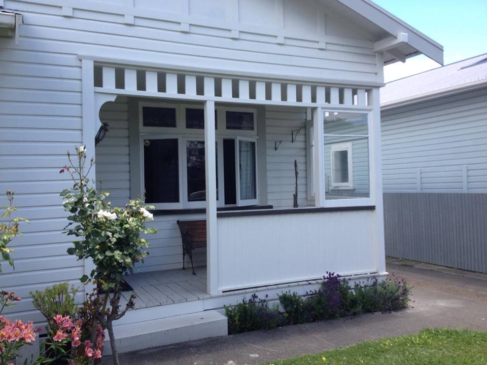 The exterior of the house features a welcoming porch with decorative wooden details and a seating area. Lush greenery and flowering plants frame the entrance, adding a touch of nature. Large windows allow natural light to brighten the space, while a gentle facade reflects classic design.