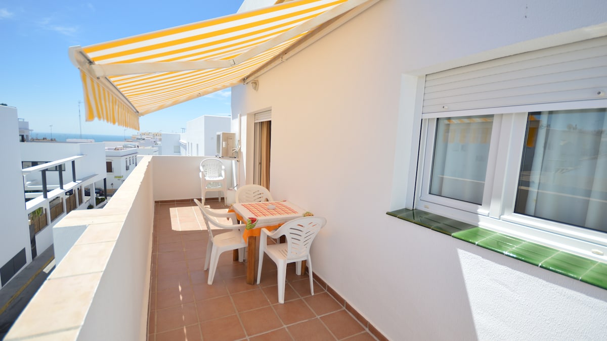 A private terrace is featured, equipped with a sunshade, a table, and four chairs, inviting outdoor dining opportunities. The view extends to the ocean, and natural light fills the space, complemented by a light-colored tiled floor.