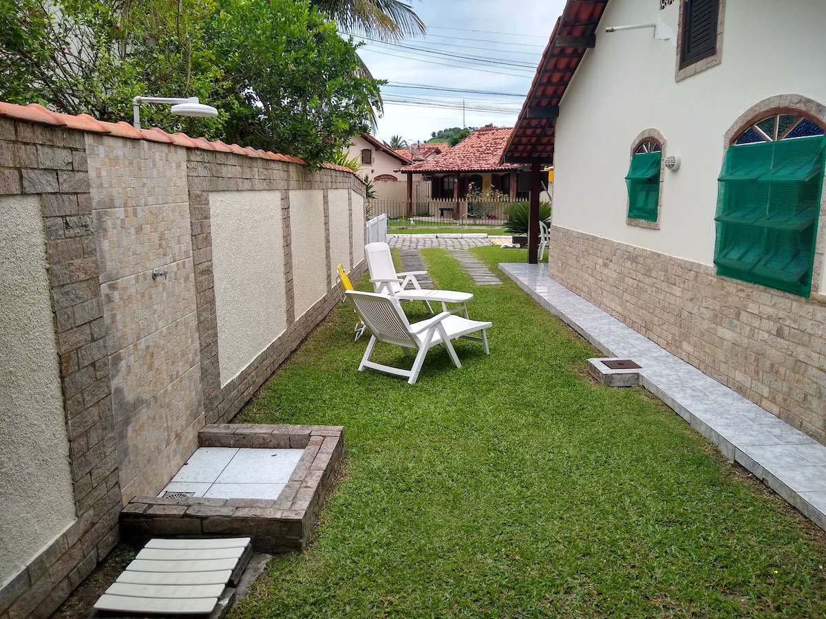 A grassy pathway is lined with two comfortable lounge chairs facing a stone shower area. The space is bordered by stone walls and features a clean, tiled pathway leading to the house, providing a serene outdoor environment for relaxation.