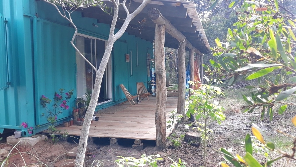 A rustic deck is shown built from natural wood, supported by tree trunks, providing a shaded area. Two wooden chairs are positioned in a relaxed manner, with colorful flowers and plants visible in the surrounding natural landscape.