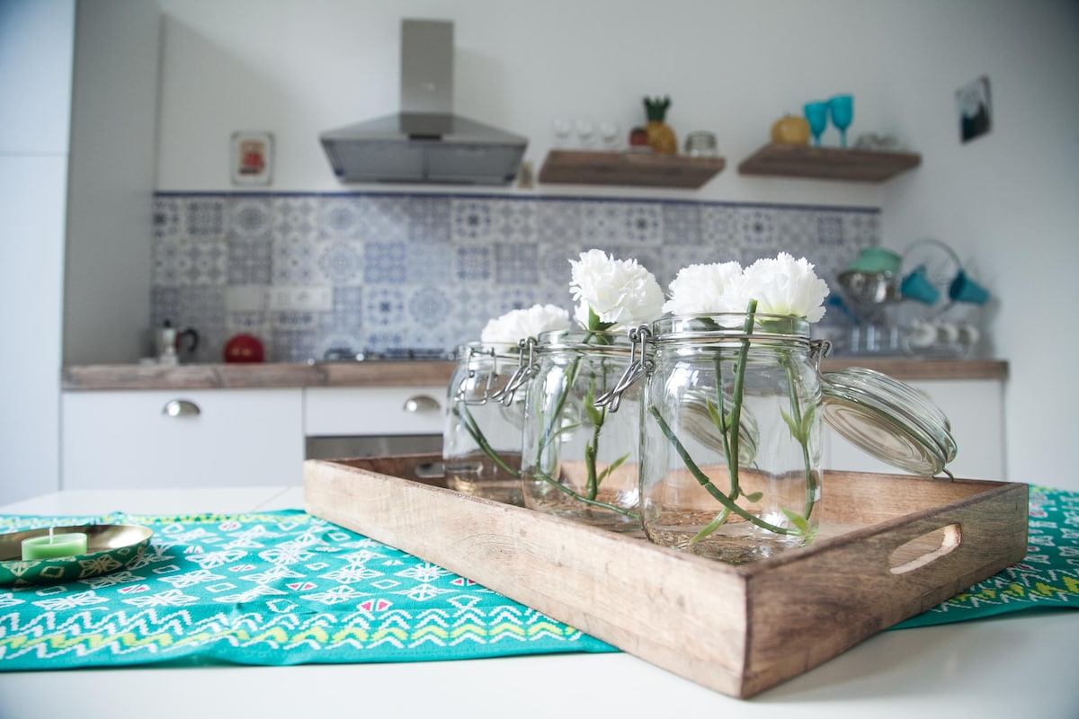 A wooden tray holds three glass jars filled with white flowers, positioned on a colorful patterned table runner. The background features a kitchen with blue tiled backsplash and open shelves displaying dishware and decorative items.