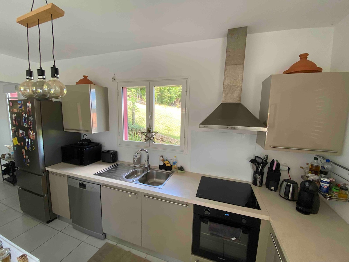 The kitchen is equipped with modern appliances, including a refrigerator, oven, and induction cooktop. Light-colored cabinetry provides ample storage. A window offers natural light and views of the outside. A circular table stands nearby for casual dining.