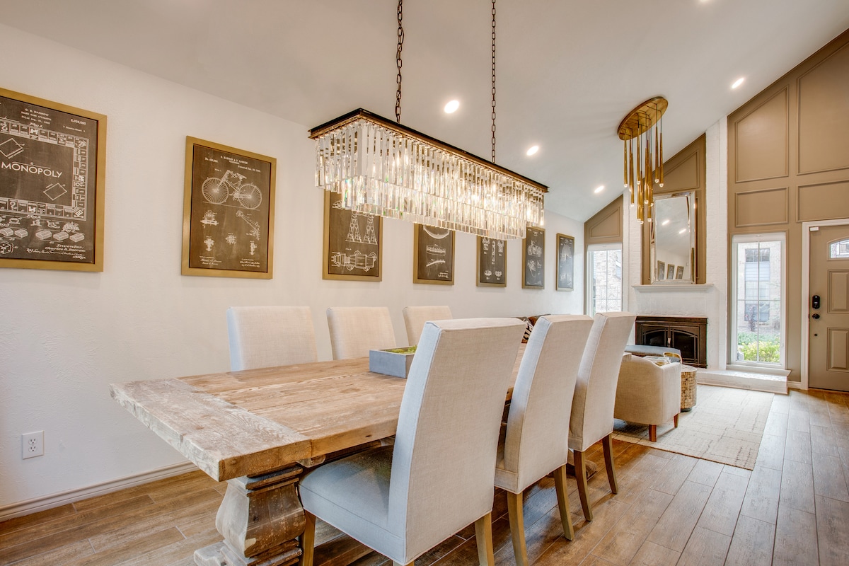 A spacious dining area features a large wooden table surrounded by upholstered chairs. Crystal chandeliers illuminate the room, complemented by art pieces hanging on light-colored walls. A fireplace presents an inviting focal point, with natural light filtering through nearby windows.