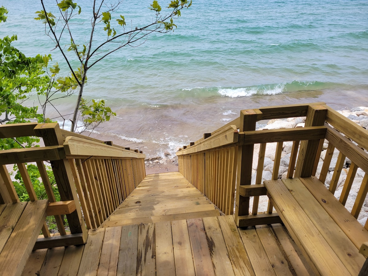 A wooden staircase leads down to a sandy shore, bordered by gentle waves lapping at the water's edge. Lush greenery frames the scene, with a view of the lake extending beyond the steps.