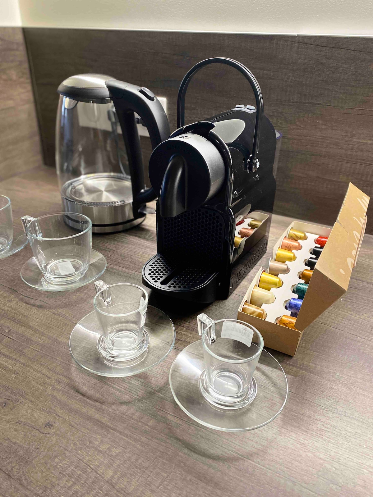 A coffee station is presented, featuring a sleek espresso machine alongside a kettle. Two glass cups with saucers are arranged in front of the machine, which is accompanied by a box of colorful coffee capsules, neatly displayed.