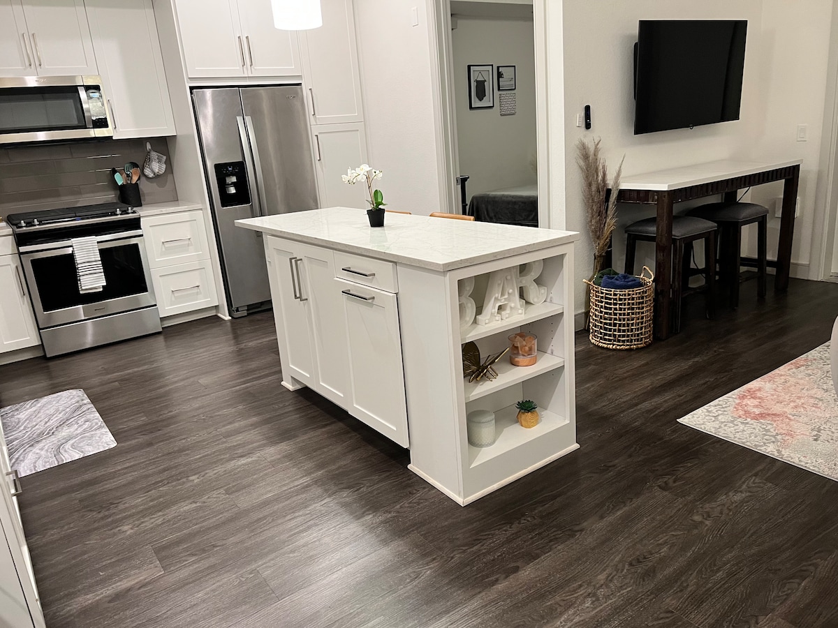 A modern kitchen is showcased with a central island topped with white stone. Stainless steel appliances, including an oven and refrigerator, complement the white cabinetry. A dining area with tall stools for four is visible near the entrance. Natural light filters through nearby rooms.