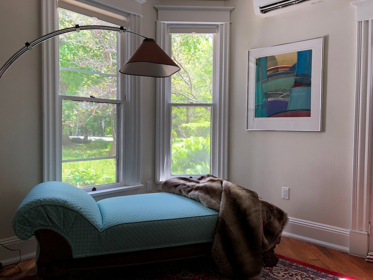 A cozy lounge area features a light blue chaise lounge, adorned with a soft throw blanket in earth tones. Large windows invite natural light while offering views of the greenery outside. A vibrant piece of artwork is displayed on the wall, enhancing the calm atmosphere.