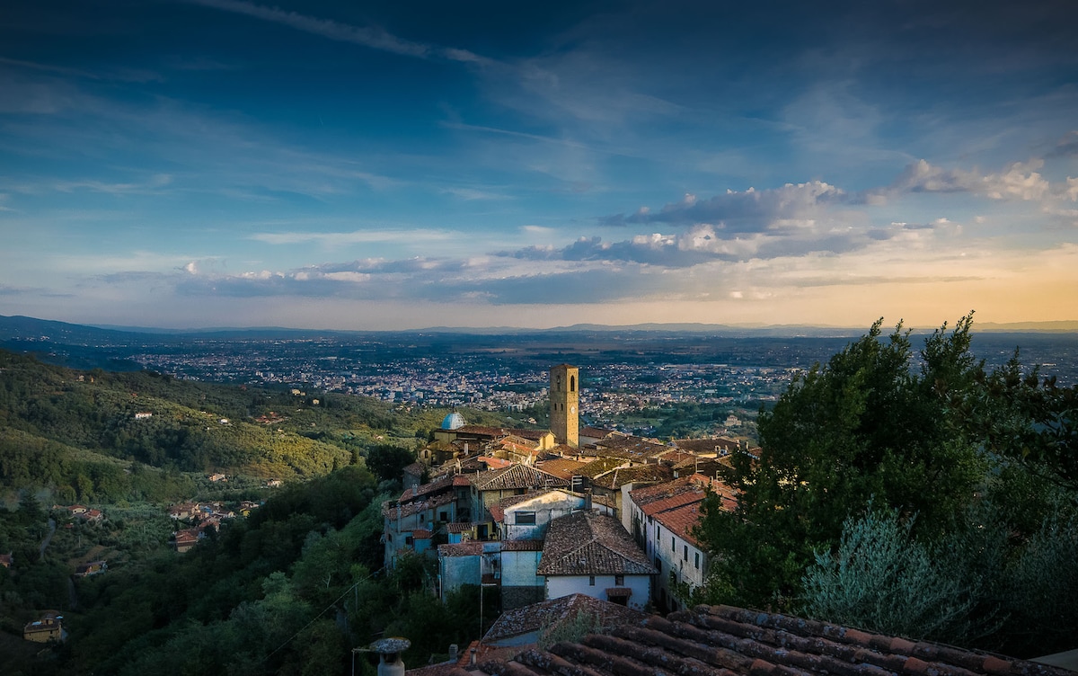 A panoramic view showcases the charming village of Massa, with its stone buildings and a prominent tower. The landscape is characterized by rolling hills and valleys, extending towards the horizon. The sky displays varying shades of blue and hints of sunset, contributing to the scenic atmosphere.