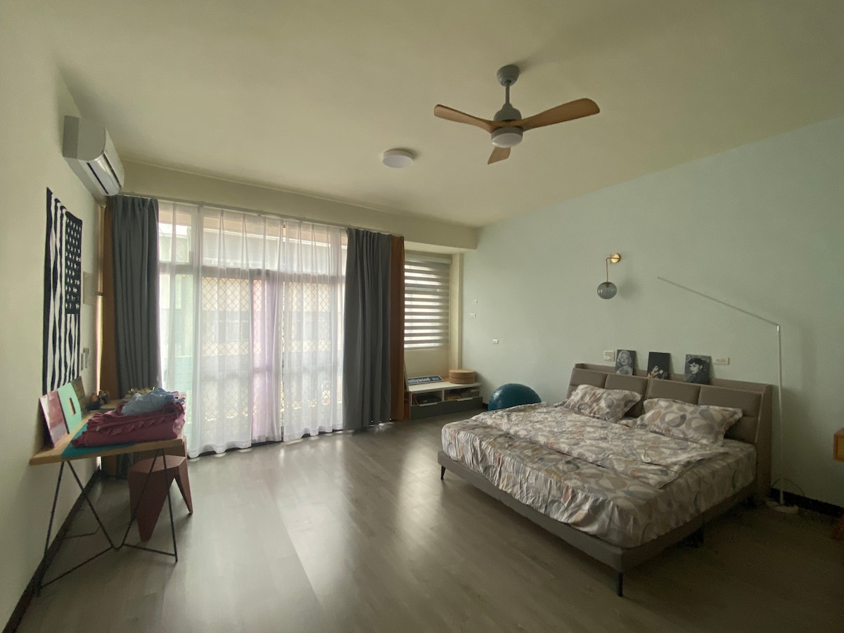 A spacious room features a queen-size bed with a light-colored comforter. Natural light filters through large windows with layered curtains, while a ceiling fan adds ventilation. A small table and a wooden bench are positioned against the wall, enhancing the room's functionality.