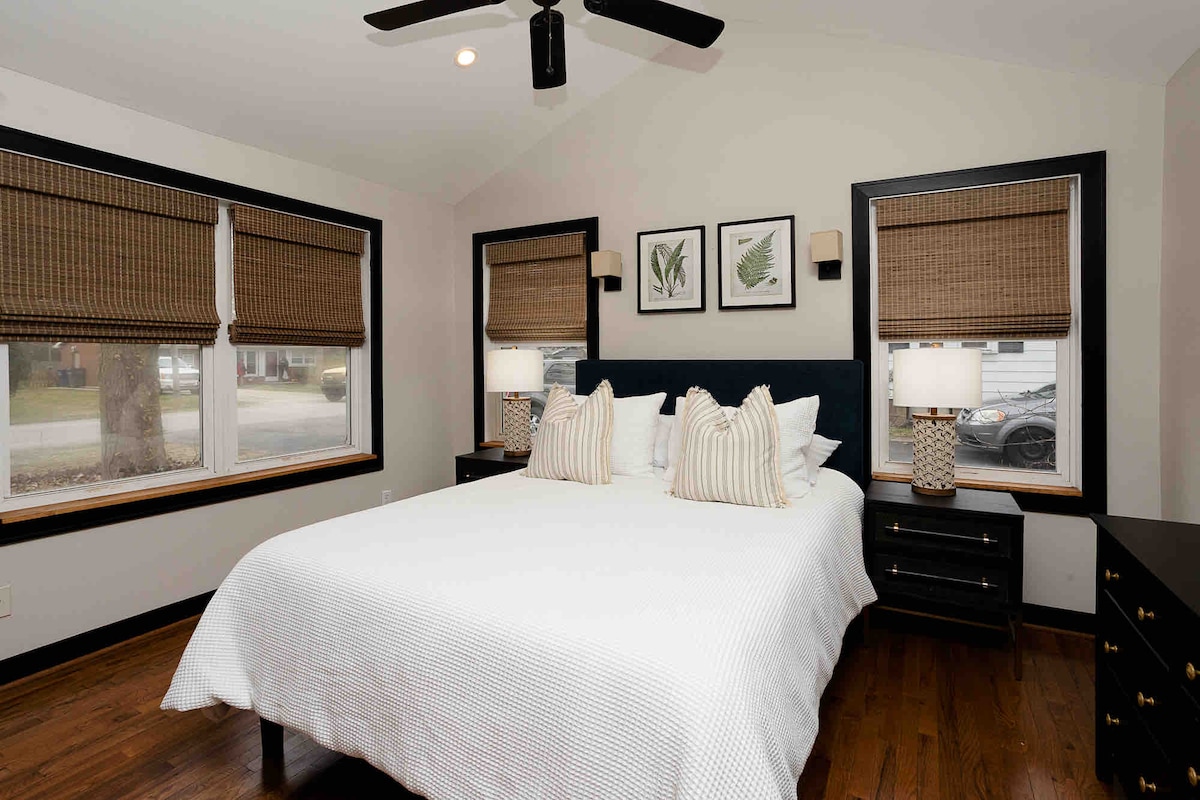 A bright bedroom features a spacious bed adorned with white linens and decorative pillows, flanked by two nightstands. Natural light enters through two windows with woven shades. Wall art depicting foliage adds a touch of nature to the cozy space.